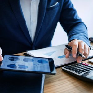 Suited men working with a calculator and tablet on top of his desk.